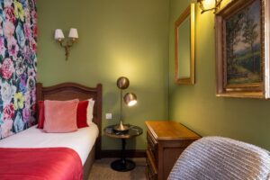 single bed in a triple room - pink pillows, green and flowers fabrics - wooden luggage rack - light - painting and chair - large room in Paris