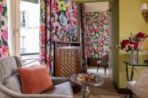 large window, flower fabrics and curtains, green fabric, grey armchair with a pink pillow, table, flowers and a corridor between the two parts of this large room Paris