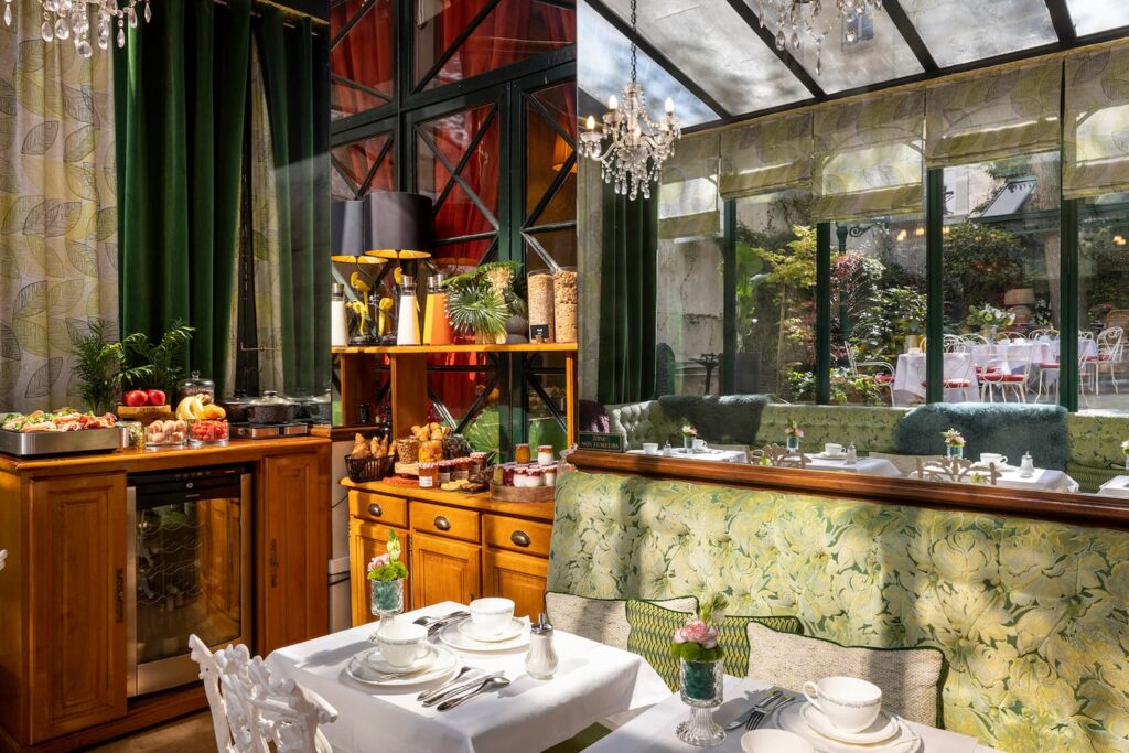 petit-déjeuner hotel paris - véranda de l'hôtel avec banquettes vertes, tables blanches et buffet dressé. Vue sur le jardin de notre hôtel de charme